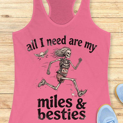 A pink racerback tank top laid flat on a light wood background, featuring a colorful graphic that reads 'all I need are my miles & besties' with a running female skeleton. Part of a blue shoe is visible at the bottom right.