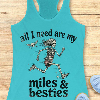 A blue racerback tank top laid flat on a light wood background, featuring a colorful graphic that reads 'all I need are my miles & besties' with a running female skeleton. Part of a blue shoe is visible at the bottom right.