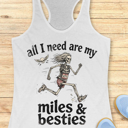 A white racerback tank top laid flat on a light wood background, featuring a colorful graphic that reads 'all I need are my miles & besties' with a running female skeleton. Part of a blue shoe is visible at the bottom right.