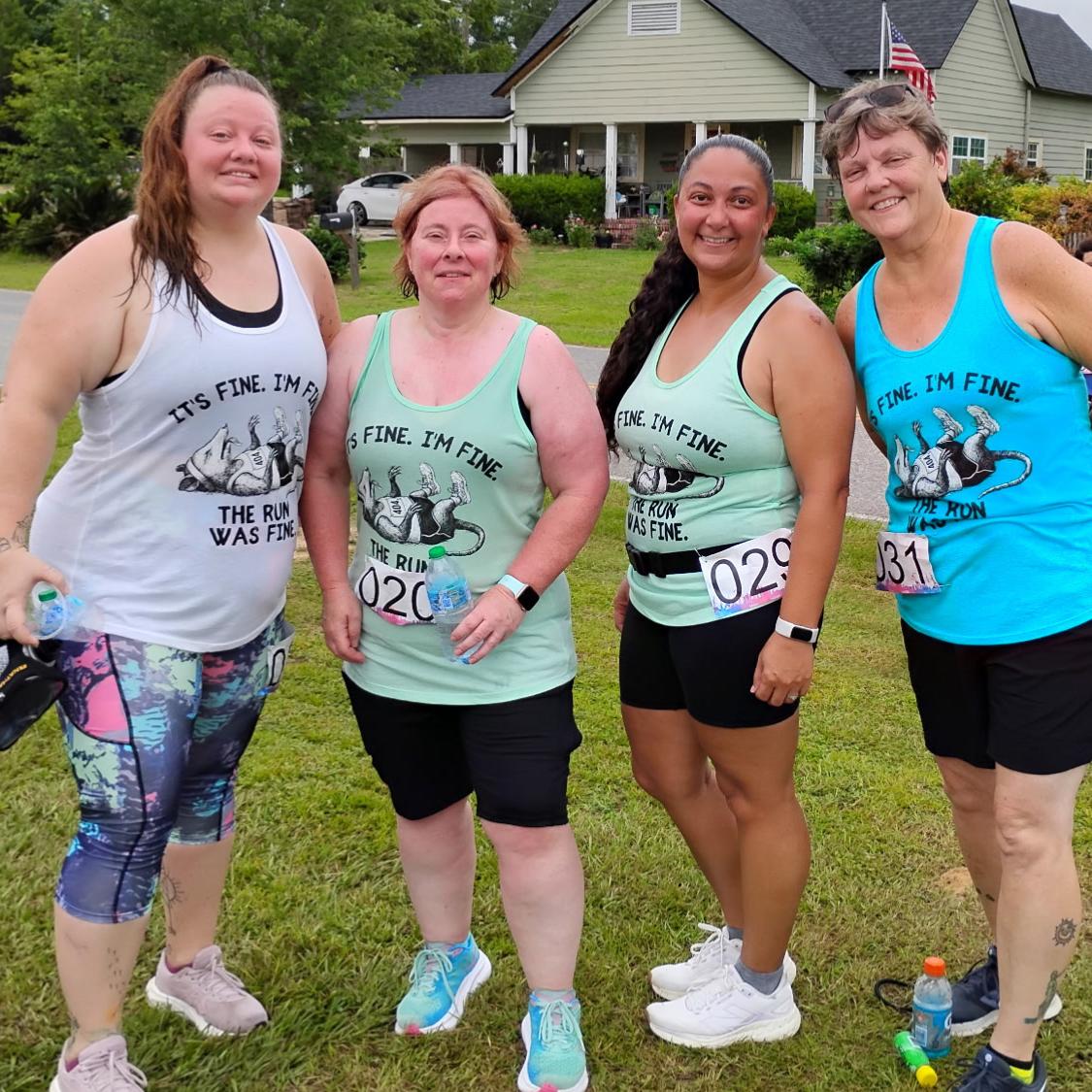 Four women standing together outdoors on a grassy lawn in front of a house, wearing matching athletic tank tops with the slogan 'IT'S FINE. I'M FINE.