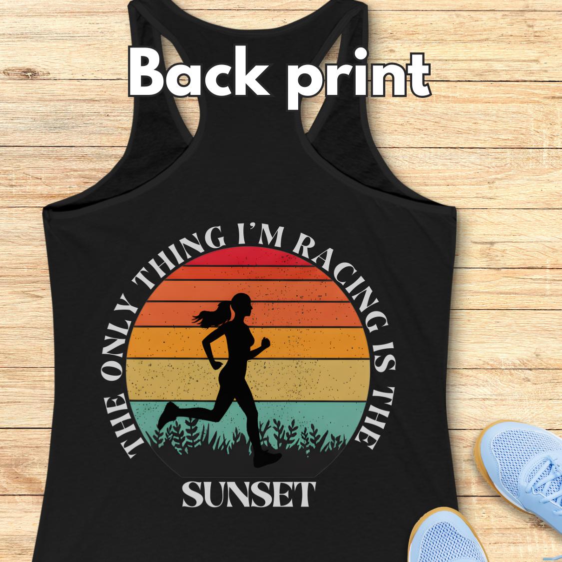 Black racerback tank top laid flat on a light wood background, with a large back print graphic showing a running silhouette over a sunset gradient and the text “The only thing I’m racing is the sunset.” White “Back print” text appears at the top, and light blue shoes are partially visible at the lower right as styling props.