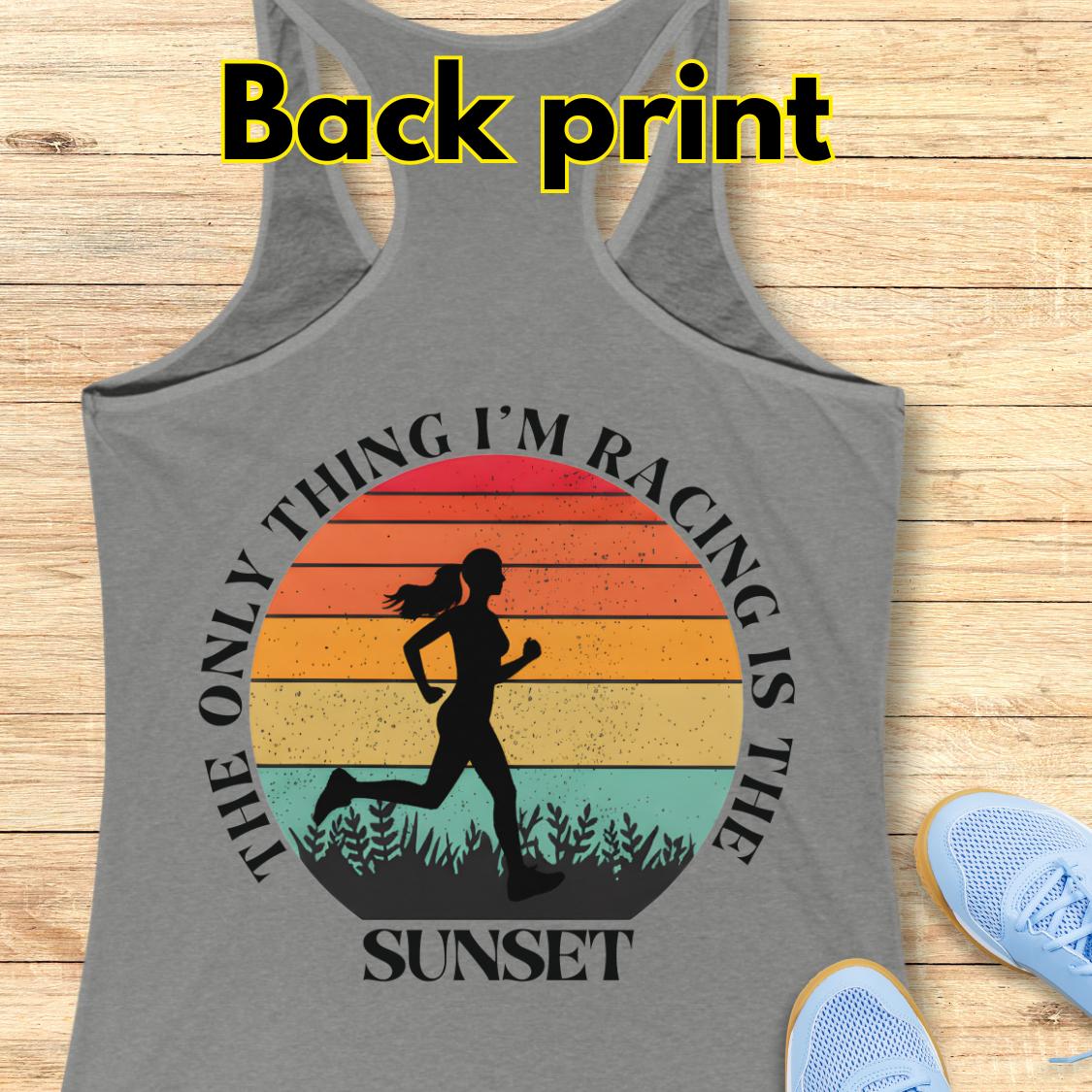 grey racerback tank top laid flat on a light wood background, with a large back print graphic showing a running silhouette over a sunset gradient and the text “The only thing I’m racing is the sunset.” White “Back print” text appears at the top, and light blue shoes are partially visible at the lower right as styling props.