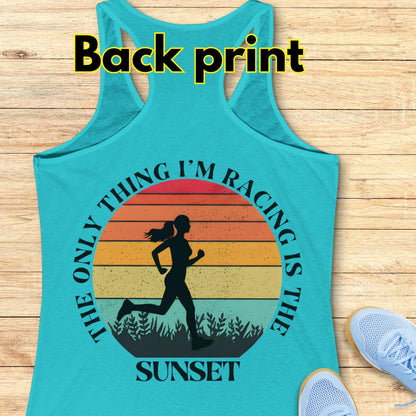 blue racerback tank top laid flat on a light wood background, with a large back print graphic showing a running silhouette over a sunset gradient and the text “The only thing I’m racing is the sunset.” White “Back print” text appears at the top, and light blue shoes are partially visible at the lower right as styling props.