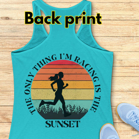 blue racerback tank top laid flat on a light wood background, with a large back print graphic showing a running silhouette over a sunset gradient and the text “The only thing I’m racing is the sunset.” White “Back print” text appears at the top, and light blue shoes are partially visible at the lower right as styling props.