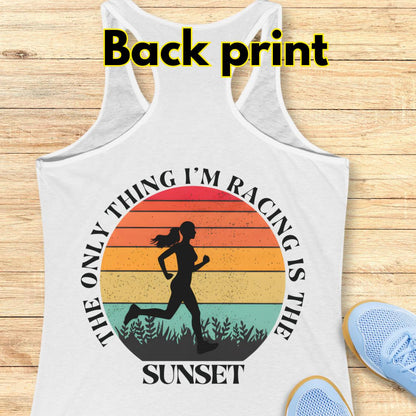 A white racerback tank top laid flat on a light wood surface, with a large back print graphic showing a running silhouette inside a sunset circle and the text 'THE ONLY THING I'M RACING IS THE SUNSET.' The words 'Back print' appear at the top, and part of a blue shoe is visible at the lower right.