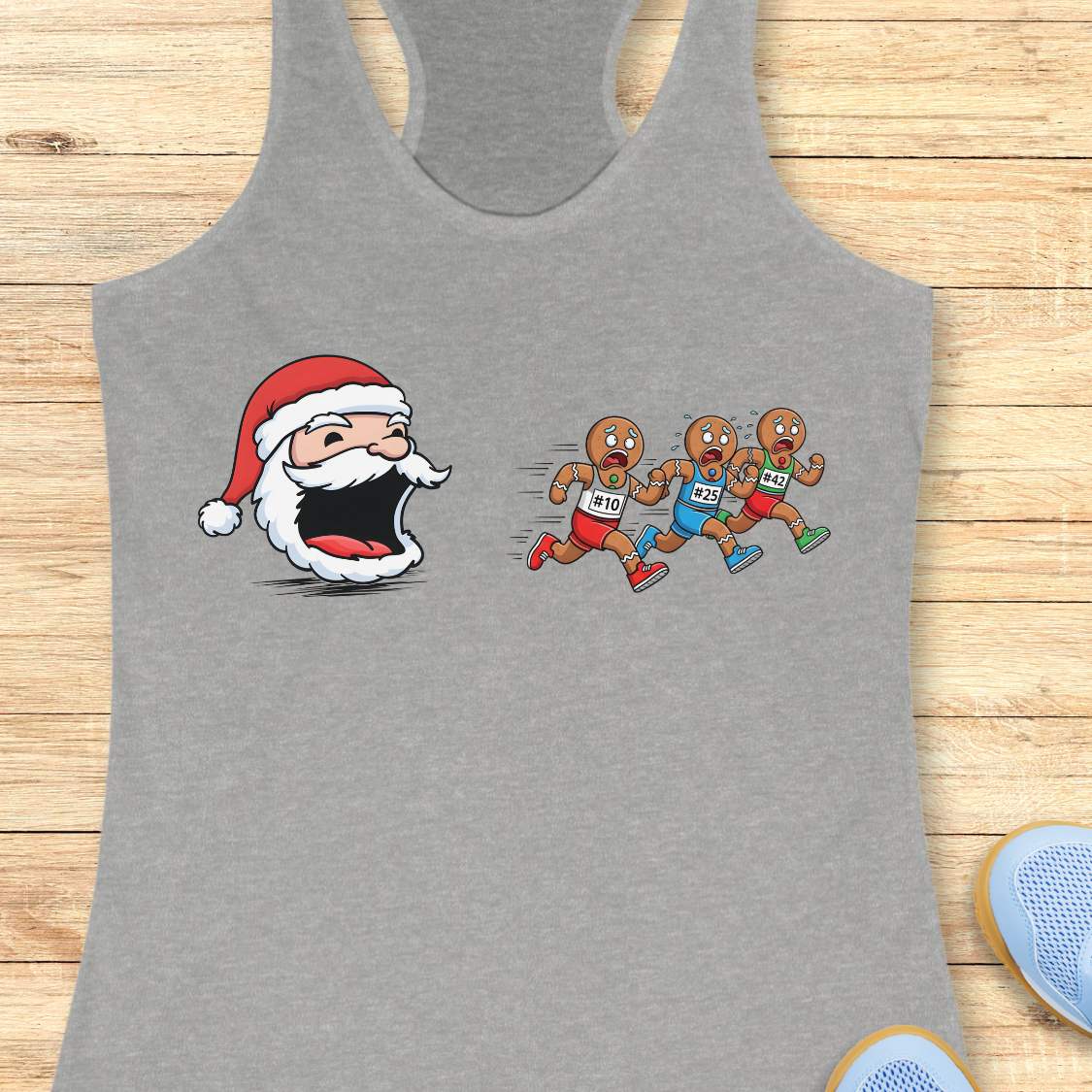 Grey tank top featuring a cartoon Santa head with a surprised expression chasing three gingerbread men wearing red blue and green shorts with race bibs #10 #25 #42 on a wooden background with white earbuds and a blue athletic shoe