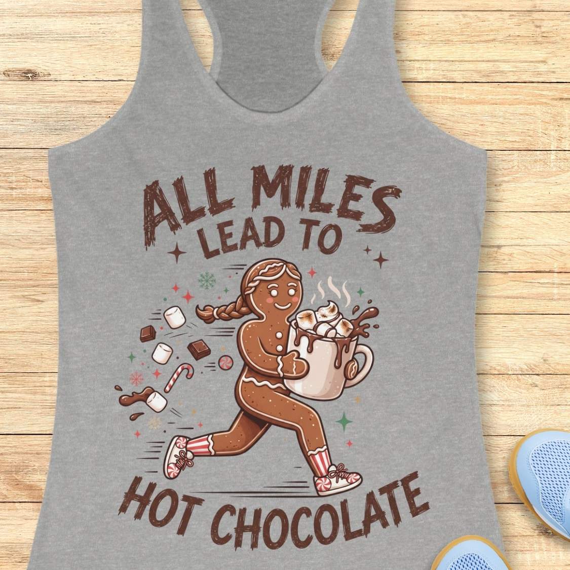 grey running tank top featuring a cartoon gingerbread runner carrying a steaming mug of hot chocolate topped with marshmallows. Chocolate pieces, marshmallows, candy canes, and winter-themed accents surround the character. The design includes the phrase 'ALL MILES LEAD TO HOT CHOCOLATE.