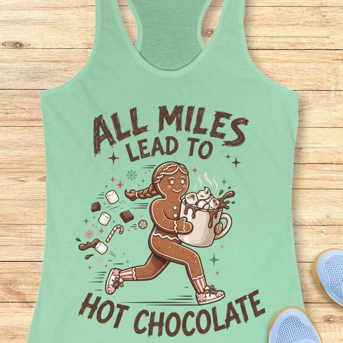 Mint green running tank top featuring a cartoon gingerbread runner carrying a steaming mug of hot chocolate topped with marshmallows. Chocolate pieces, marshmallows, candy canes, and winter-themed accents surround the character. The design includes the phrase 'ALL MILES LEAD TO HOT CHOCOLATE.
