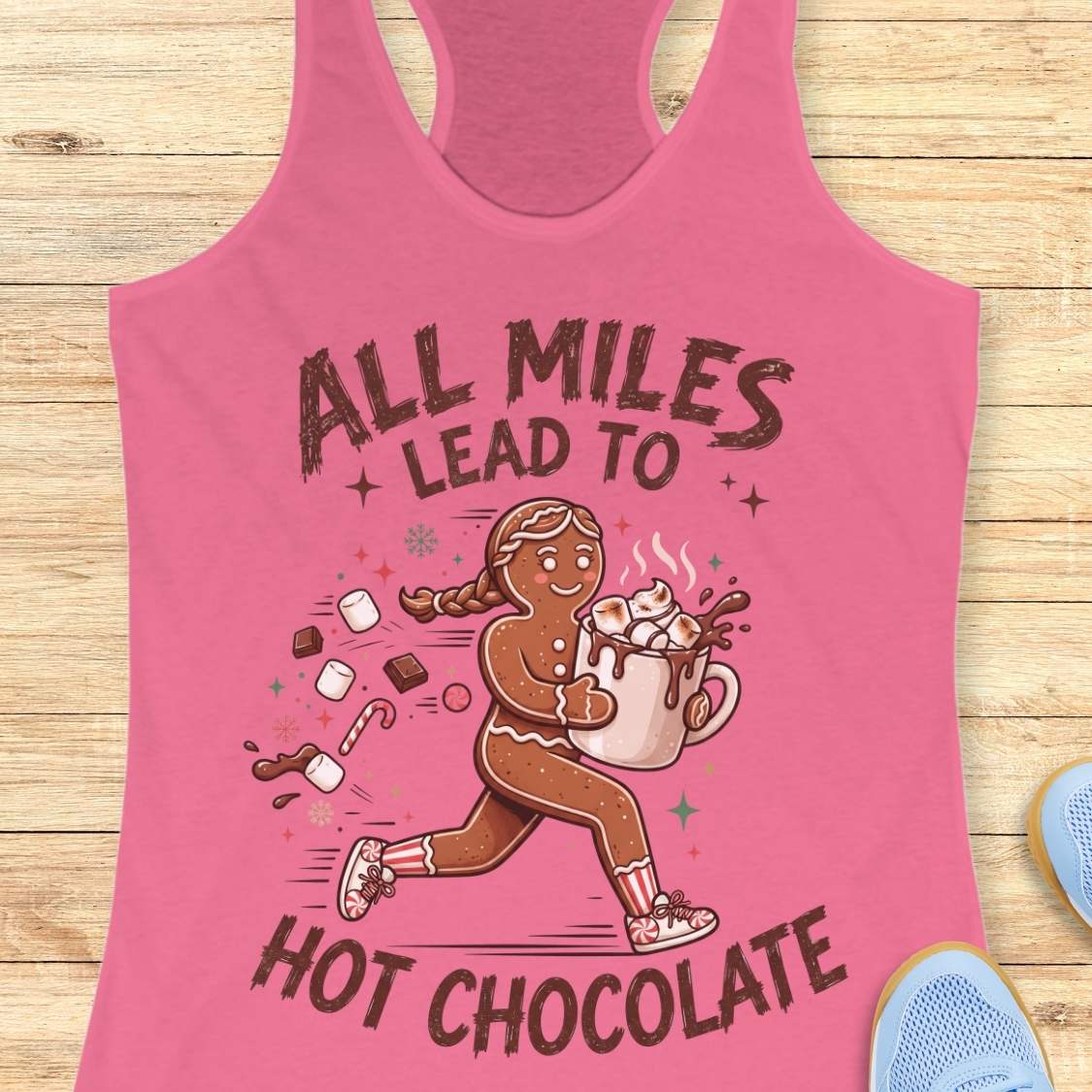 pink running tank top featuring a cartoon gingerbread runner carrying a steaming mug of hot chocolate topped with marshmallows. Chocolate pieces, marshmallows, candy canes, and winter-themed accents surround the character. The design includes the phrase 'ALL MILES LEAD TO HOT CHOCOLATE.