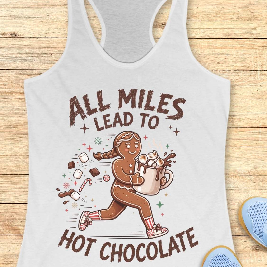 white running tank top featuring a cartoon gingerbread runner carrying a steaming mug of hot chocolate topped with marshmallows. Chocolate pieces, marshmallows, candy canes, and winter-themed accents surround the character. The design includes the phrase 'ALL MILES LEAD TO HOT CHOCOLATE.
