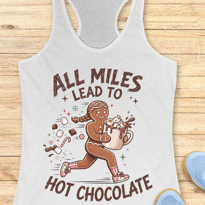 white running tank top featuring a cartoon gingerbread runner carrying a steaming mug of hot chocolate topped with marshmallows. Chocolate pieces, marshmallows, candy canes, and winter-themed accents surround the character. The design includes the phrase 'ALL MILES LEAD TO HOT CHOCOLATE.
