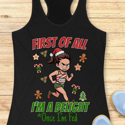 black women's racerback tank top featuring a cartoon holiday runner in a striped red, green, and white running outfit with a Santa hat. The runner is surrounded by festive Christmas elements including candy canes, gingerbread cookies, holly, star cookies, and a Christmas tree. The design reads: “FIRST OF ALL” above the runner and “I’M A DELIGHT Once I’m Fed” below. The artwork combines holiday humor with running culture in a playful, festive style.