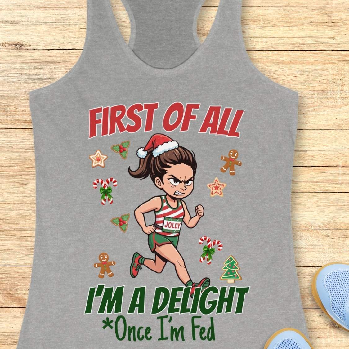 heather grey women's racerback tank top featuring a cartoon holiday runner in a striped red, green, and white running outfit with a Santa hat. The runner is surrounded by festive Christmas elements including candy canes, gingerbread cookies, holly, star cookies, and a Christmas tree. The design reads: “FIRST OF ALL” above the runner and “I’M A DELIGHT Once I’m Fed” below. The artwork combines holiday humor with running culture in a playful, festive style.