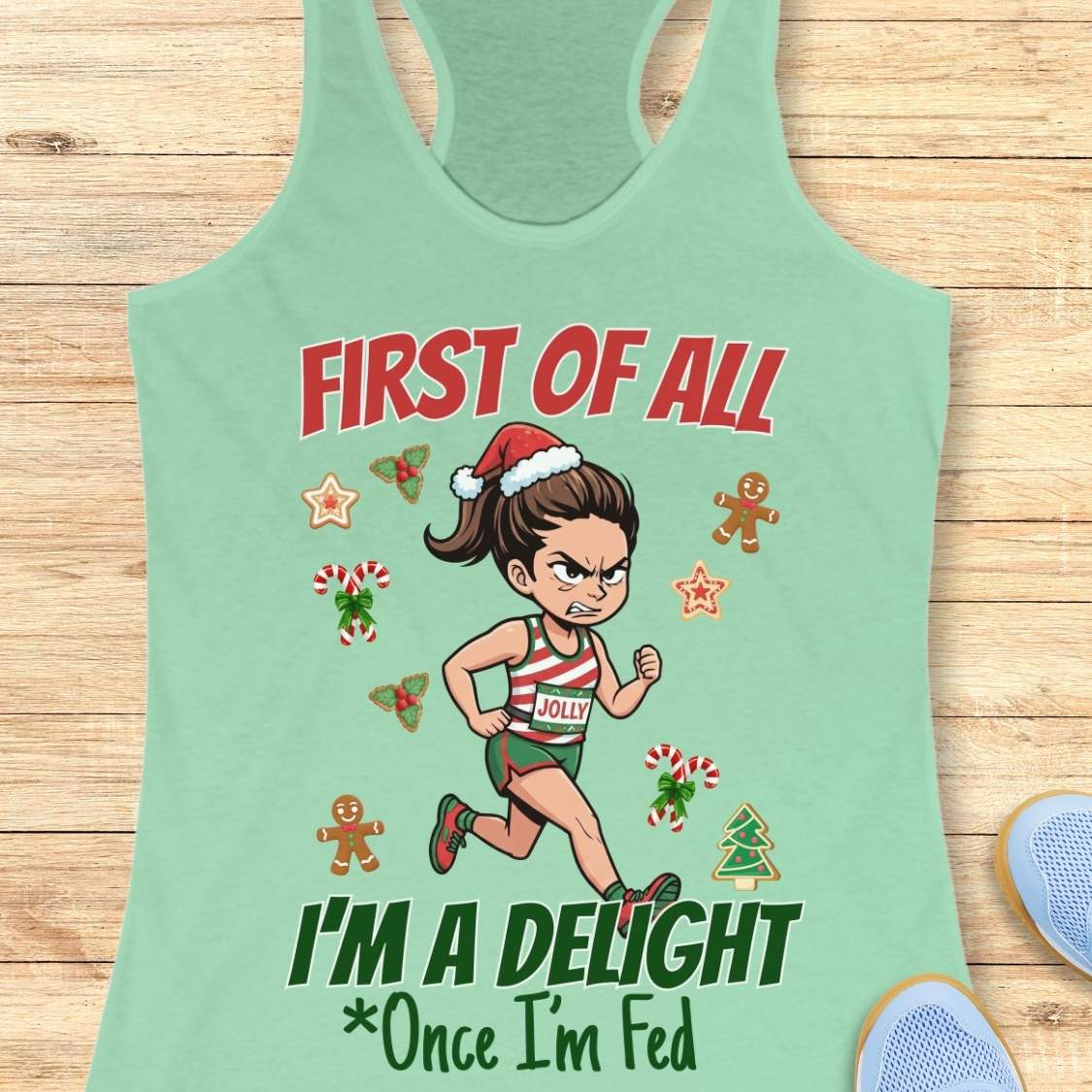 Mint green women's racerback tank top featuring a cartoon holiday runner in a striped red, green, and white running outfit with a Santa hat. The runner is surrounded by festive Christmas elements including candy canes, gingerbread cookies, holly, star cookies, and a Christmas tree. The design reads: “FIRST OF ALL” above the runner and “I’M A DELIGHT Once I’m Fed” below. The artwork combines holiday humor with running culture in a playful, festive style.