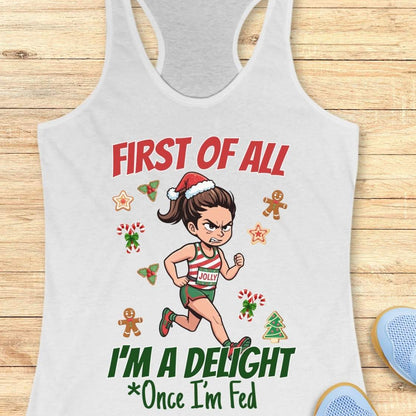 white women's racerback tank top featuring a cartoon holiday runner in a striped red, green, and white running outfit with a Santa hat. The runner is surrounded by festive Christmas elements including candy canes, gingerbread cookies, holly, star cookies, and a Christmas tree. The design reads: “FIRST OF ALL” above the runner and “I’M A DELIGHT Once I’m Fed” below. The artwork combines holiday humor with running culture in a playful, festive style.