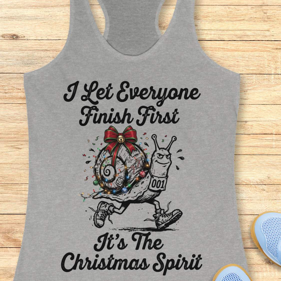 A heather gray racerback tank top laid flat on a light wood background, featuring a graphic of a decorated snail running with the text 'I Let Everyone Finish First It's The Christmas Spirit'.