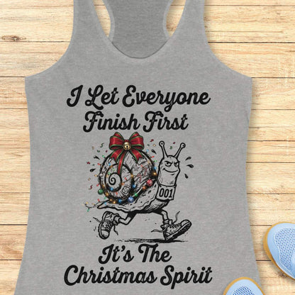 A heather gray racerback tank top laid flat on a light wood background, featuring a graphic of a decorated snail running with the text 'I Let Everyone Finish First It's The Christmas Spirit'.