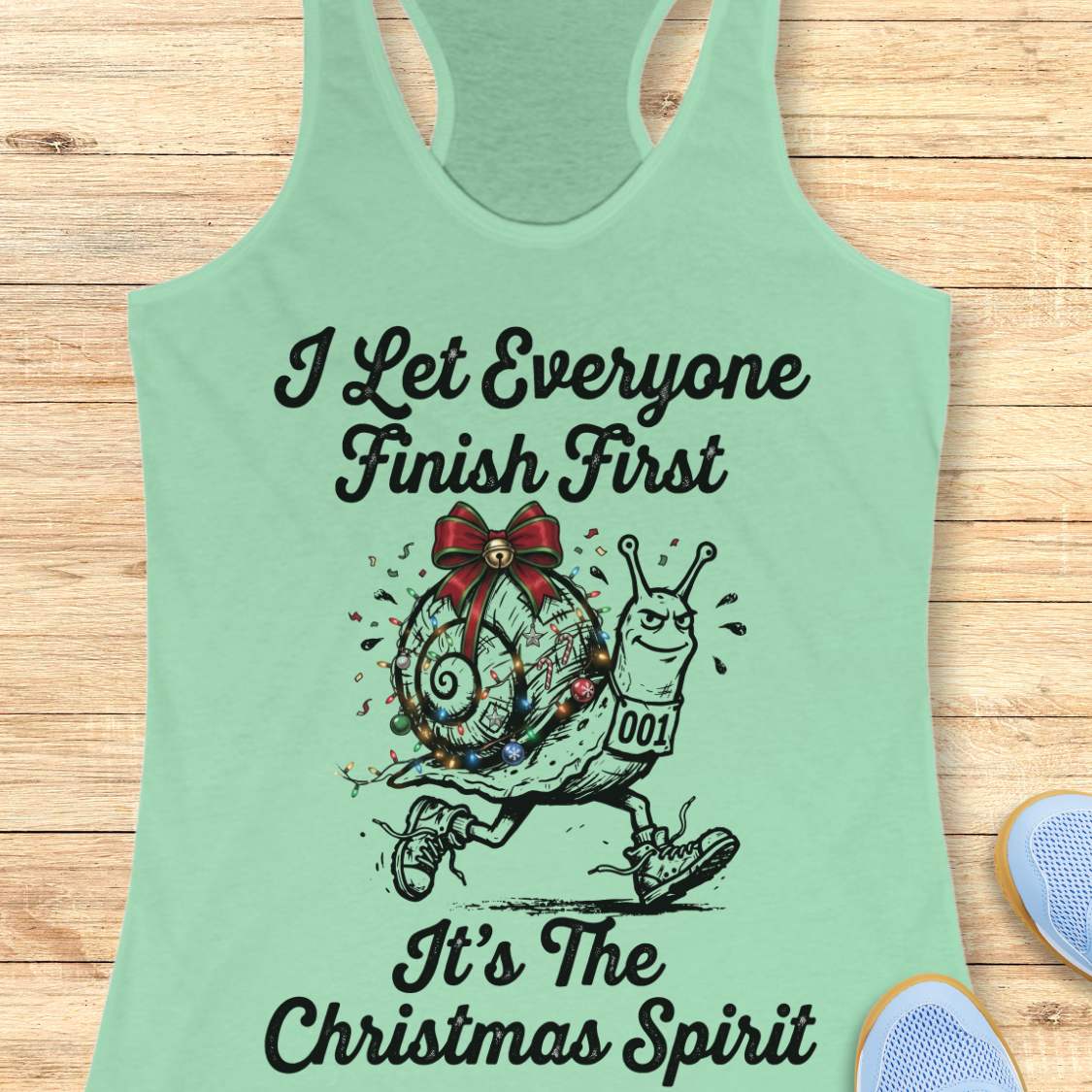 A mint green racerback tank top laid flat on a light wood background, featuring a black graphic of a decorated snail running with the text 'I Let Everyone Finish First It's The Christmas Spirit'.