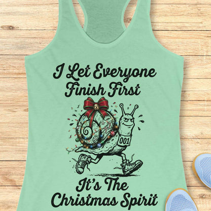 A mint green racerback tank top laid flat on a light wood background, featuring a black graphic of a decorated snail running with the text 'I Let Everyone Finish First It's The Christmas Spirit'.