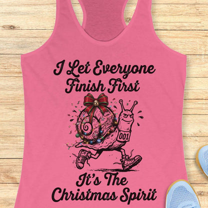 A pink racerback tank top laid flat on a light wood surface, featuring a black graphic of a snail carrying a decorated Christmas ornament with the text 'I Let Everyone Finish First It's The Christmas Spirit.' Partial blue shoes are visible at the edges as styling props.