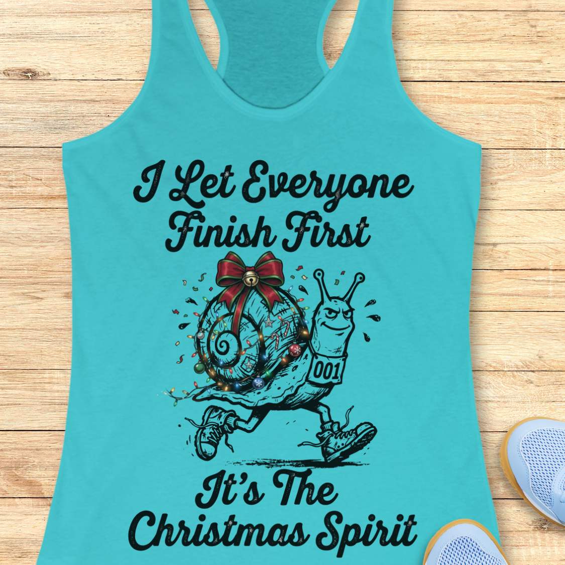 A blue racerback tank top laid flat on a light wood background, featuring a black graphic of a decorated snail running with the text 'I Let Everyone Finish First It's The Christmas Spirit'.