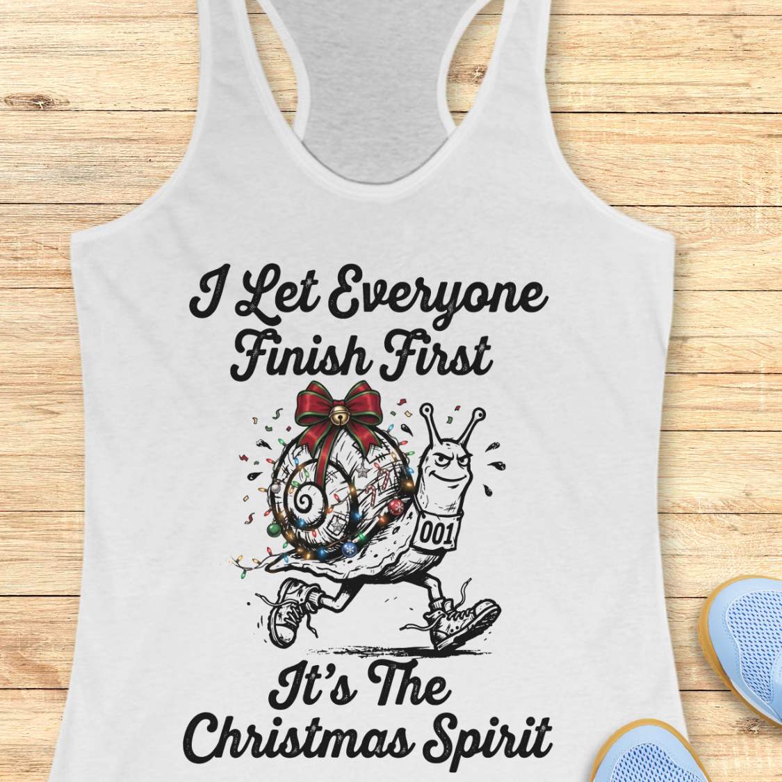 A white racerback tank top laid flat on a light wood background, featuring a black graphic of a snail wearing a red bow and multicolored Christmas lights, with the text 'I Let Everyone Finish First It's The Christmas Spirit'.