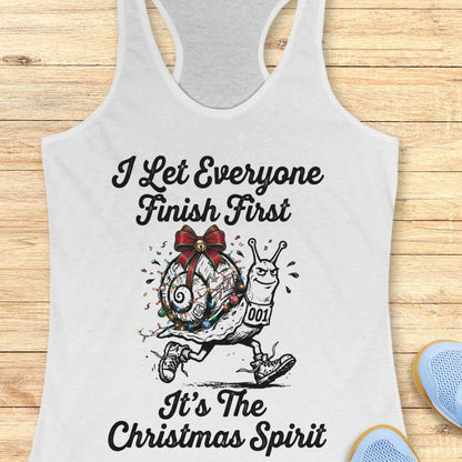 A white racerback tank top laid flat on a light wood background, featuring a black graphic of a snail wearing a red bow and multicolored Christmas lights, with the text 'I Let Everyone Finish First It's The Christmas Spirit'.