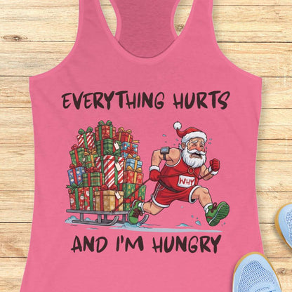 A pink racerback tank top laid flat on a light wood background, featuring a graphic of a Santa Claus runner pushing a sleigh filled with Christmas presents. The text reads 'EVERYTHING HURTS AND I'M HUNGRY' in black lettering.