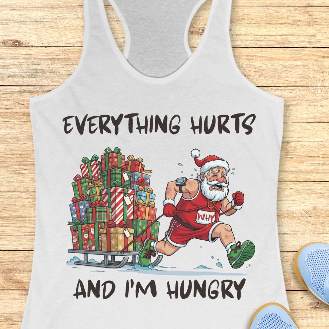 A white racerback tank top laid flat on a light wood surface, featuring a festive graphic of Santa running while pulling a sled full of Christmas presents. The text reads 'EVERYTHING HURTS AND I'M HUNGRY'.