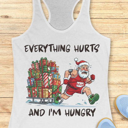 A white racerback tank top laid flat on a light wood surface, featuring a festive graphic of Santa running while pulling a sled full of Christmas presents. The text reads 'EVERYTHING HURTS AND I'M HUNGRY'.