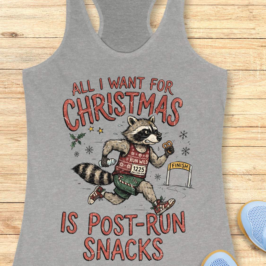 A grey racerback tank top laid flat on a light wood surface, featuring a humorous graphic of a raccoon running a 5K with the text 'ALL I WANT FOR CHRISTMAS IS POST-RUN SNACKS'. A blue shoe is partially visible in the bottom right corner as a styling prop.