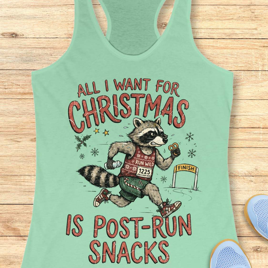 A mint green racerback tank top laid flat on a light wood background, featuring a humorous graphic of a raccoon running like a marathon finish line with the text 'ALL I WANT FOR CHRISTMAS IS POST-RUN SNACKS'.