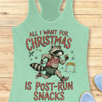 A mint green racerback tank top laid flat on a light wood background, featuring a humorous graphic of a raccoon running like a marathon finish line with the text 'ALL I WANT FOR CHRISTMAS IS POST-RUN SNACKS'.