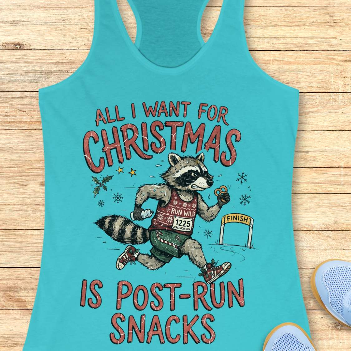 A turquoise racerback tank top laid flat on a light wood surface, featuring a humorous graphic of a raccoon running a 5K with the text 'ALL I WANT FOR CHRISTMAS IS POST-RUN SNACKS'. A blue shoe is partially visible in the bottom right corner as a styling prop.