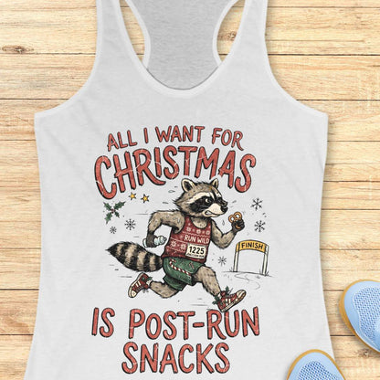 A white racerback tank top laid flat on a light wood surface, featuring a humorous graphic that reads 'ALL I WANT FOR CHRISTMAS IS POST-RUN SNACKS' around an illustrated raccoon running with Christmas-themed snacks. Part of a blue shoe is visible at the bottom right.