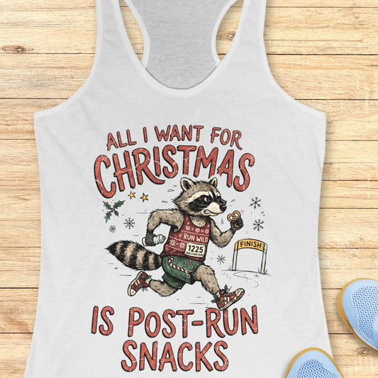 A white racerback tank top laid flat on a light wood surface, featuring a humorous graphic that reads 'ALL I WANT FOR CHRISTMAS IS POST-RUN SNACKS' around an illustrated raccoon running with Christmas-themed snacks. Part of a blue shoe is visible at the bottom right.