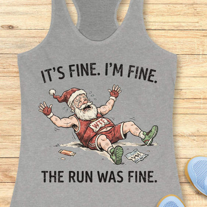 A heather gray racerback tank top laid flat on a light wood background, featuring a humorous graphic of a cartoon Santa running and falling over with the text 'IT'S FINE. I'M FINE.