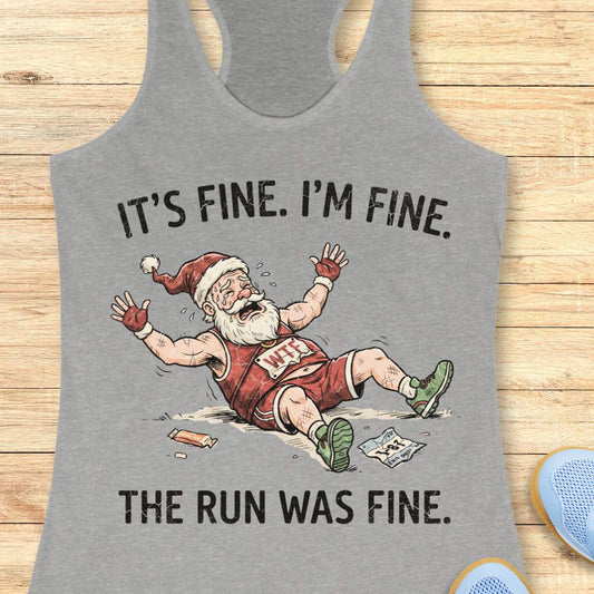 A heather gray racerback tank top laid flat on a light wood background, featuring a humorous graphic of a cartoon Santa running and falling over with the text 'IT'S FINE. I'M FINE.
