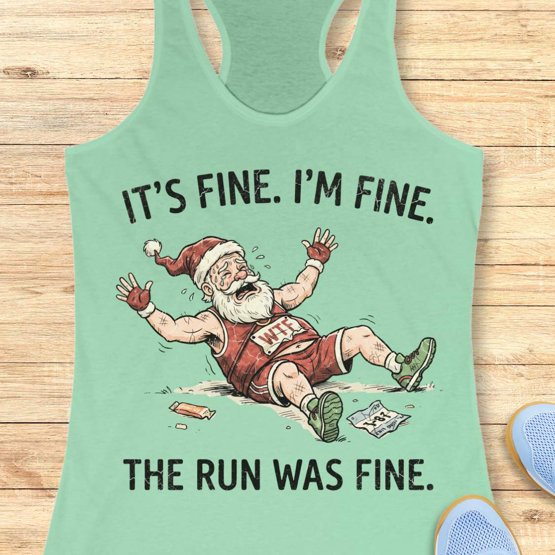 A mint green racerback tank top laid flat on a light wood background, featuring a humorous graphic of a distressed Santa Claus in a red running outfit with the text 'IT'S FINE. I'M FINE.
