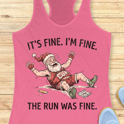 A pink racerback tank top laid flat on a light wood surface, featuring a humorous graphic of a cartoon Santa Claus in a red and white basketball uniform lying on the ground with the text 'IT'S FINE. I'M FINE.