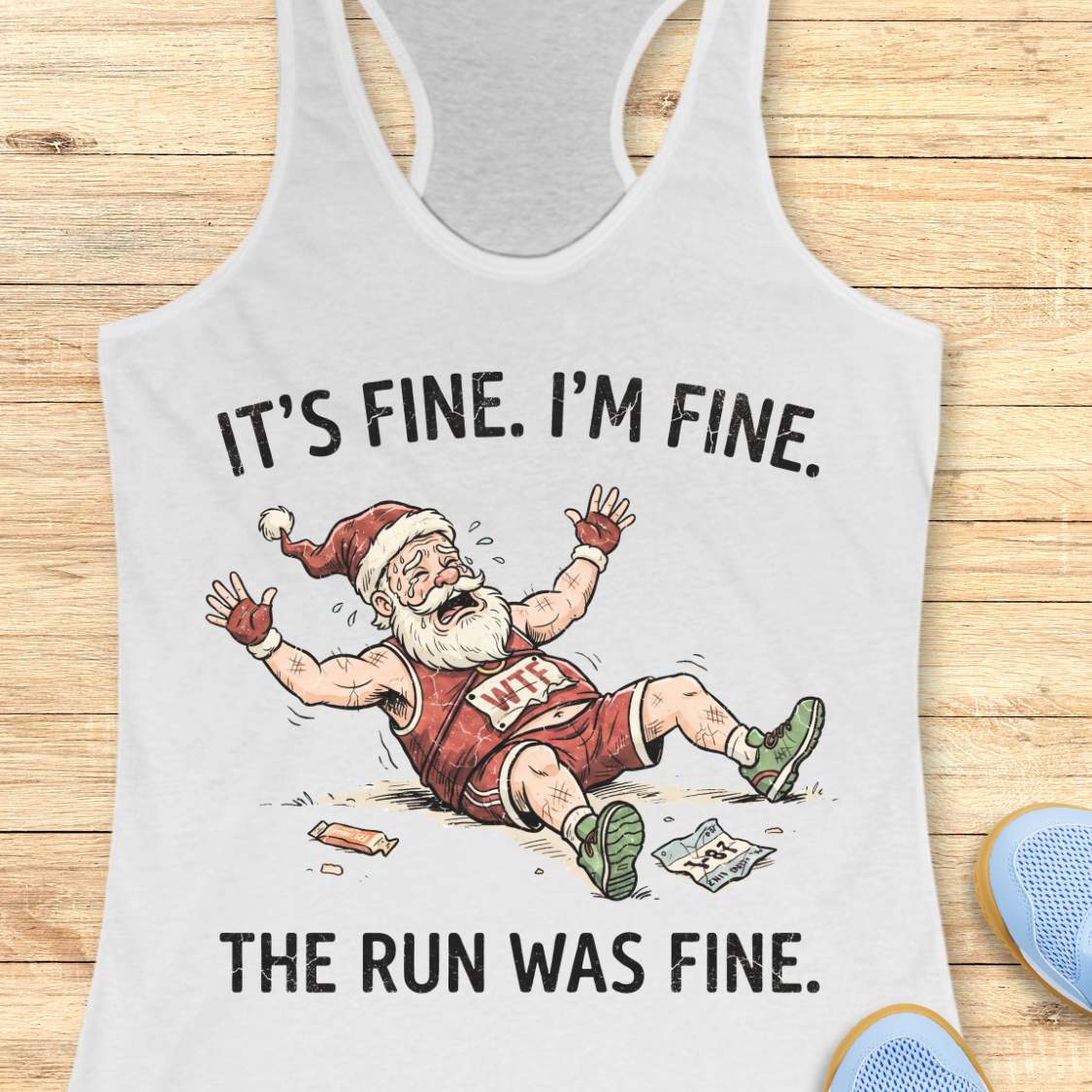 A white racerback tank top laid flat on a light wood background, featuring a humorous graphic of a disappointed Santa Claus running with a broken leg. The text reads 'IT'S FINE.