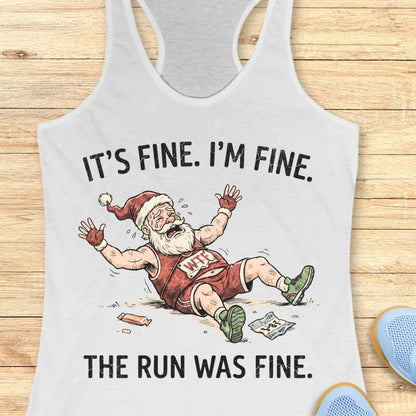 A white racerback tank top laid flat on a light wood background, featuring a humorous graphic of a disappointed Santa Claus running with a broken leg. The text reads 'IT'S FINE.
