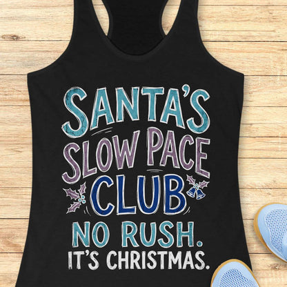 A black racerback tank top laid flat on a light wood background, featuring the text 'SANTA'S SLOW PACE CLUB NO RUSH. IT'S CHRISTMAS.
