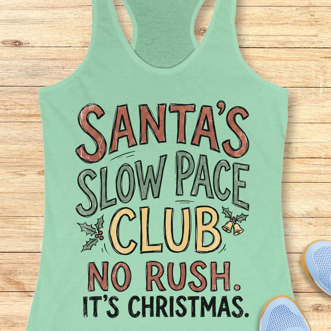 A mint green racerback tank top laid flat on a light wood background, featuring a festive graphic that reads 'SANTA'S SLOW PACE CLUB NO RUSH. IT'S CHRISTMAS.