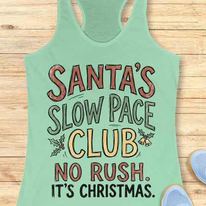 A mint green racerback tank top laid flat on a light wood background, featuring a festive graphic that reads 'SANTA'S SLOW PACE CLUB NO RUSH. IT'S CHRISTMAS.