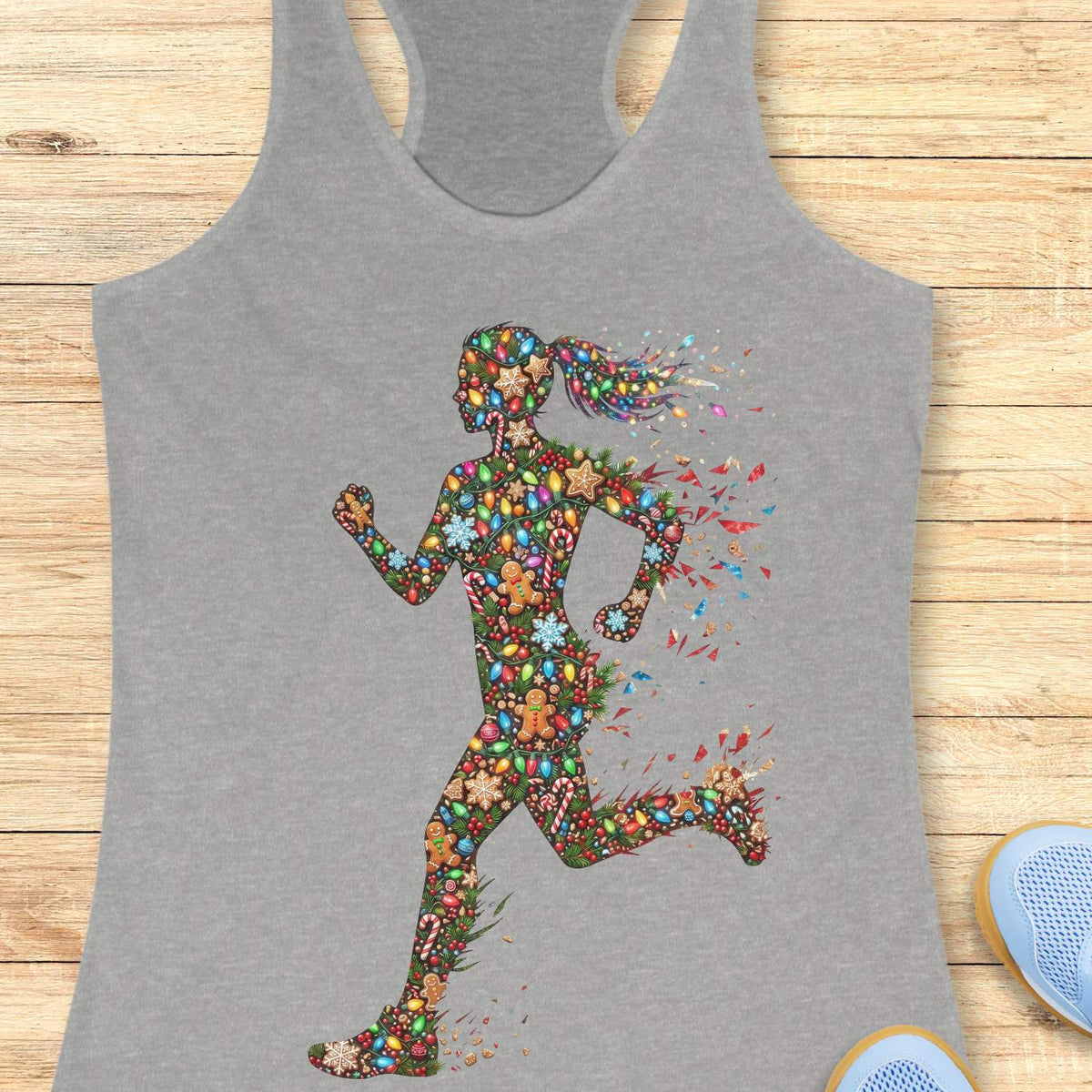 grey racerback tank top displayed on a light wood surface, featuring a colorful silhouette of a running figure made from festive Christmas elements, including string lights, gingerbread cookies, candy canes, snowflakes, ornaments, and holly. The runner's ponytail and limbs appear to dissolve into bright, scattered decorative pieces, creating a dynamic sense of motion. Partial views of blue athletic shoes are visible in the bottom corners of the image.