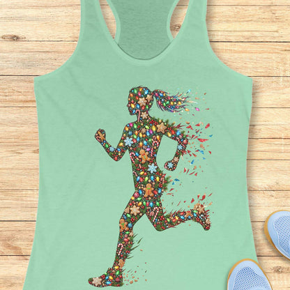Mint green racerback tank top displayed on a light wood surface, featuring a colorful silhouette of a running figure made from festive Christmas elements, including string lights, gingerbread cookies, candy canes, snowflakes, ornaments, and holly. The runner's ponytail and limbs appear to dissolve into bright, scattered decorative pieces, creating a dynamic sense of motion. Partial views of blue athletic shoes are visible in the bottom corners of the image.