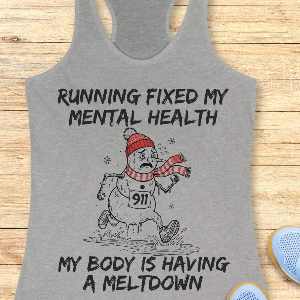 grey racerback tank top displayed on a light wooden surface featuring a humorous running-themed Christmas design. The graphic shows a melting snowman wearing a red knit hat, striped scarf, running shoes, and race bib number “911,” splashing through a puddle while running. Bold black text above and below the illustration reads: “RUNNING FIXED MY MENTAL HEALTH” and “MY BODY IS HAVING A MELTDOWN.” Partial blue athletic shoes are visible in the lower corners of the image.