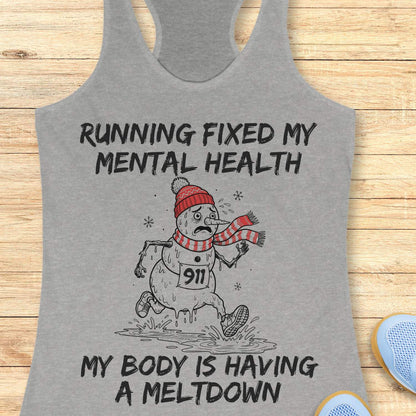 grey racerback tank top displayed on a light wooden surface featuring a humorous running-themed Christmas design. The graphic shows a melting snowman wearing a red knit hat, striped scarf, running shoes, and race bib number “911,” splashing through a puddle while running. Bold black text above and below the illustration reads: “RUNNING FIXED MY MENTAL HEALTH” and “MY BODY IS HAVING A MELTDOWN.” Partial blue athletic shoes are visible in the lower corners of the image.