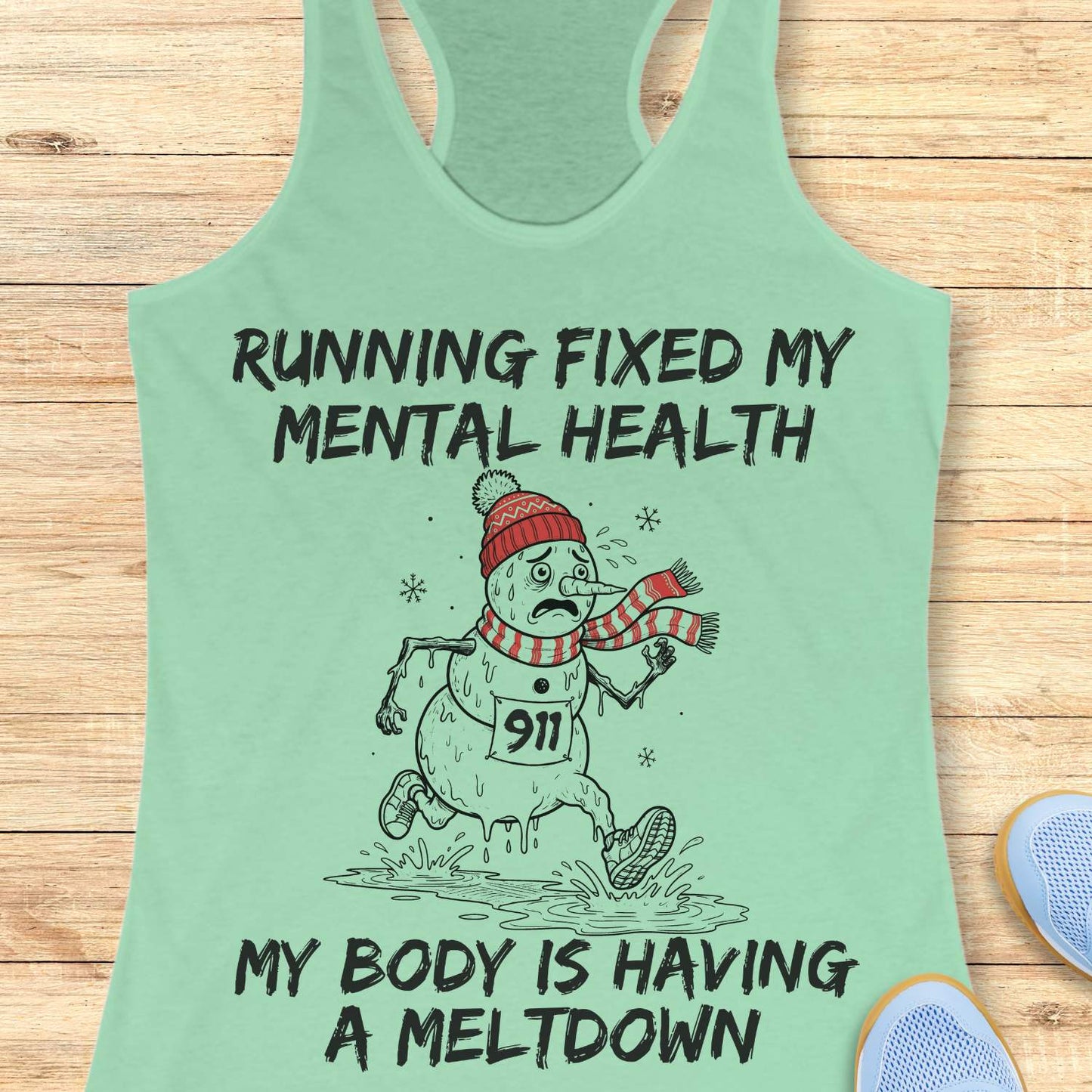 mint racerback tank top displayed on a light wooden surface featuring a humorous running-themed Christmas design. The graphic shows a melting snowman wearing a red knit hat, striped scarf, running shoes, and race bib number “911,” splashing through a puddle while running. Bold black text above and below the illustration reads: “RUNNING FIXED MY MENTAL HEALTH” and “MY BODY IS HAVING A MELTDOWN.” Partial blue athletic shoes are visible in the lower corners of the image.